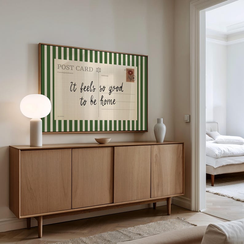 May include: A wooden sideboard with four doors and a light-colored finish. A framed postcard with green and white stripes and the text "It feels so good to be home" hangs above. A white lamp and vase are on the sideboard.