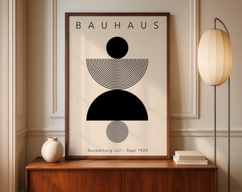 May include: A framed Bauhaus art print featuring a black geometric design on a cream background. The word "BAUHAUS" is at the top. Below the design is the text "Ausstellung Juli - Sept 1923". The print is displayed on a wooden cabinet.