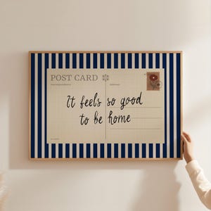 May include: A framed postcard print with the text "It feels so good to be home." The postcard design features a vintage aesthetic with a floral stamp and lined writing space. The frame has a navy blue and white striped border.