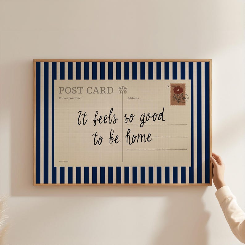 May include: A framed postcard print with the text "It feels so good to be home." The postcard design features a vintage aesthetic with a floral stamp and lined writing space. The frame has a navy blue and white striped border.