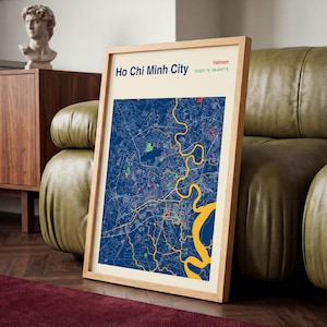 Könnte beinhalten: Ein gerahmter Karten-Druck von Ho-Chi-Minh-Stadt, Vietnam. Die Karte zeigt ein detailliertes blaues und gelbes Design mit dem Namen und den Koordinaten der Stadt. Der Druck befindet sich in einem hellen Holzrahmen und lehnt an einem grünen Ledersofa.