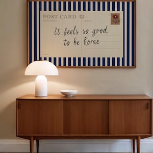 May include: A mid-century modern wooden sideboard with a white lamp and bowl on top. A framed postcard with blue stripes and the text "It feels so good to be home" hangs above the sideboard. The room has a neutral color palette.