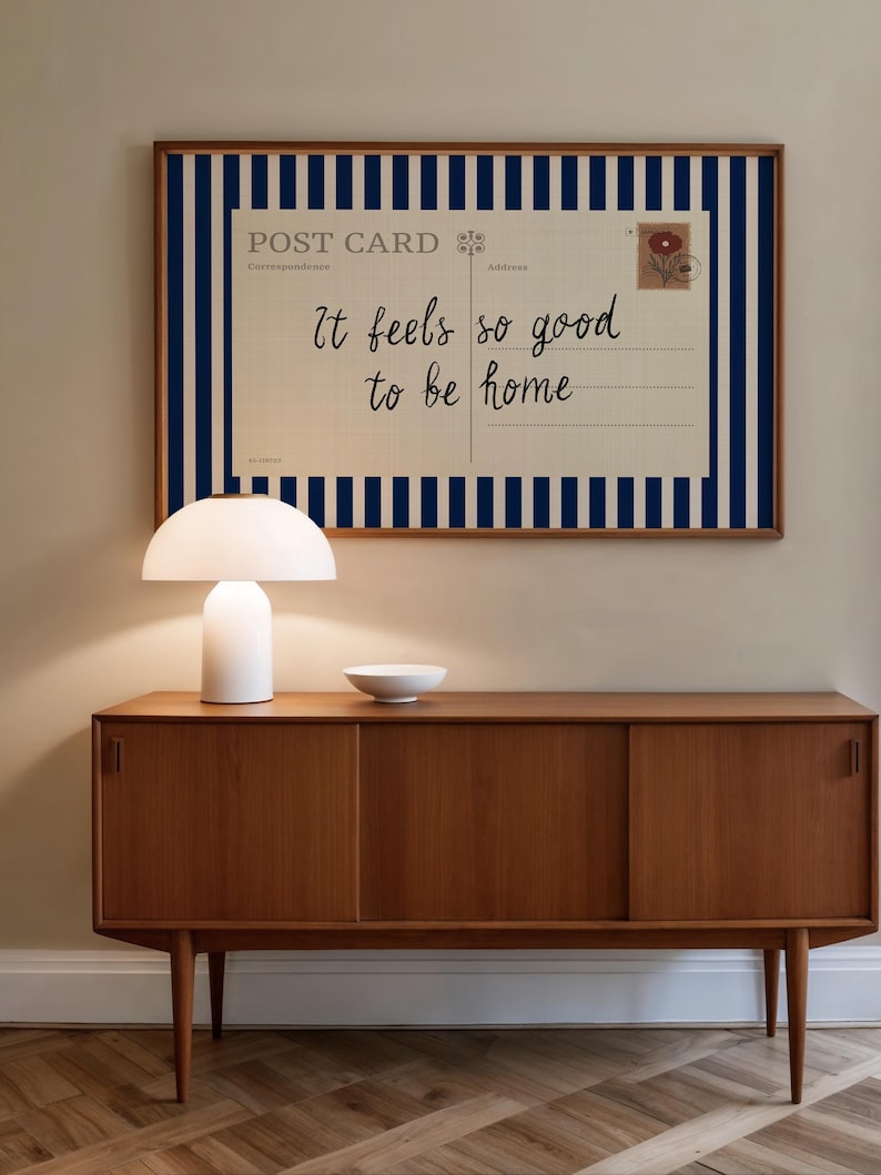 May include: A mid-century modern wooden sideboard with a white lamp and bowl on top. A framed postcard with blue stripes and the text "It feels so good to be home" hangs above the sideboard. The room has a neutral color palette.