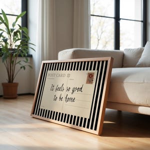 May include: A framed postcard with the text "It feels so good to be home." The postcard has a beige background with a floral stamp and is set against a black and white striped border. The frame is a light wood color.