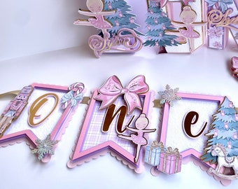 May include: Pink and gold 'One' banner with a ballerina, a Christmas tree, and a nutcracker. The banner is decorated with bows, snowflakes, and candy canes. The banner also includes the name 'Sylvie'.