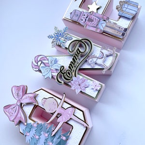 Könnte beinhalten: Rosa und goldfarbene dekorative Boxen in Form des Buchstabens "E", verziert mit Weihnachts- und Ballettthemen. Mit Nussknacker, Schneeflocke, Zuckerstange, Schaukelpferd und Ballerina. Der Name "Emma" ist ebenfalls enthalten.