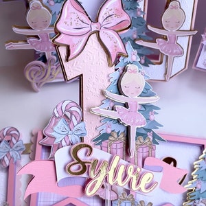 Op de afbeelding: Een roze en gouden taarttopper met een ballerina, een kerstboom, cadeautjes en een snoepstok. De topper is versierd met glitter en heeft de naam "Sylvie" erop.