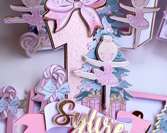 May include: A pink and gold cake topper featuring a ballerina, a Christmas tree, presents, and a candy cane. The topper is decorated with glitter and has the name "Sylvie" written on it.