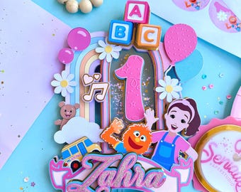 Può includere: Un cake topper colorato con il nome "Zahra" in glitter rosa, con un numero uno, personaggi dei cartoni animati e un arcobaleno. Include blocchi ABC, palloncini e dettagli di margherite. Il tema è una festa di primo compleanno.