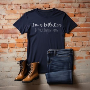 May include: A navy blue t-shirt with the text "I'm a reflection of your intentions" in white lettering. The shirt is paired with blue jeans and brown leather boots.