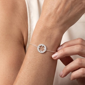 May include: A delicate silver-toned chain bracelet featuring a circular charm with a cut-out four-leaf clover design. The bracelet is secured with a thin metal chain that wraps around the wrist, showcasing a polished metallic finish throughout.