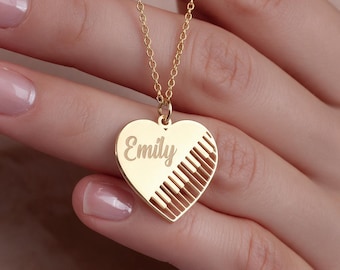 May include: A gold-toned heart-shaped pendant necklace featuring the name Emily engraved in a cursive script on the left side and a cutout design of piano keys on the right side, suspended from a delicate matching gold-toned chain link necklace.