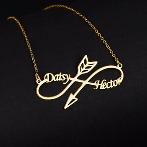 May include: Gold necklace with an infinity symbol and an arrow through it. The names "Daisy" and "Hector" are written on the infinity symbol.