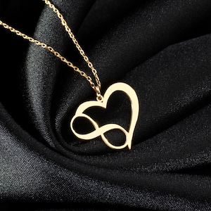 Könnte beinhalten: Eine goldene Halskette mit einem herzförmigen Anhänger, der ein Unendlichkeitszeichen trägt. Die Halskette wird auf einem schwarzen Stoffhintergrund präsentiert.