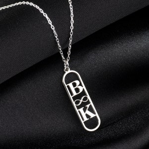 May include: A silver-colored necklace with a pendant featuring the letters "B" and "K" separated by an infinity symbol. The pendant is a long, rectangular shape with rounded ends. The necklace chain is delicate and silver.