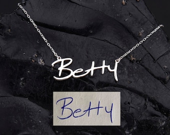 May include: A silver necklace with the name "Betty" written in cursive script. The necklace is on a black background with a handwritten note that says "Betty" on a white piece of paper.