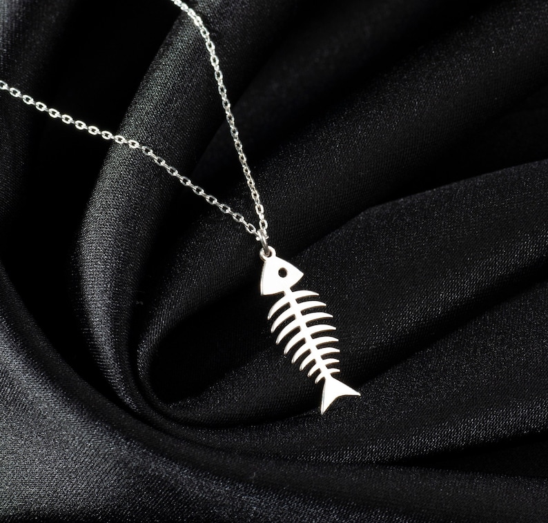 May include: A silver necklace with a fishbone pendant on a black fabric background.