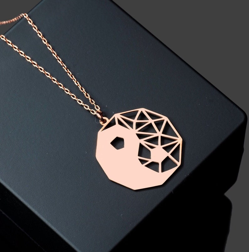 May include: A rose gold necklace with a geometric yin yang pendant. The pendant is made of metal and has a cut-out design.