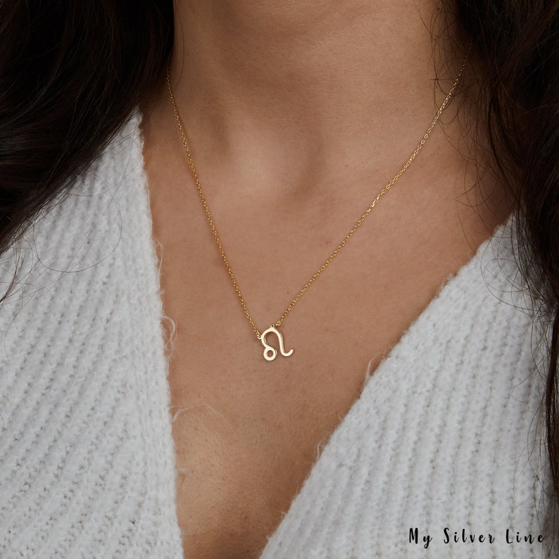 May include: A gold chain necklace with a small, delicate Leo zodiac sign pendant. The necklace is worn by a person with long brown hair.