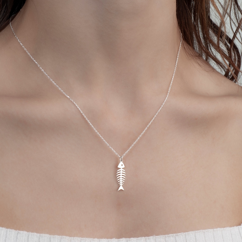 May include: A silver necklace with a delicate fish bone pendant. The necklace is on a model wearing a white shirt.