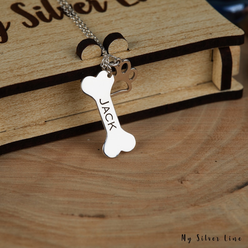 May include: Silver necklace with a bone charm engraved with the name "JACK" and a paw print charm. The necklace is displayed in a wooden box.