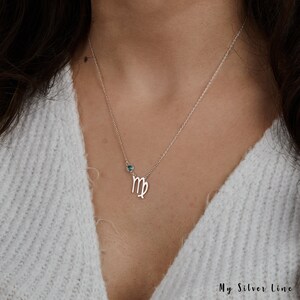 May include: Silver necklace with a Virgo zodiac symbol charm and a blue gemstone. The necklace is on a model wearing a white sweater.