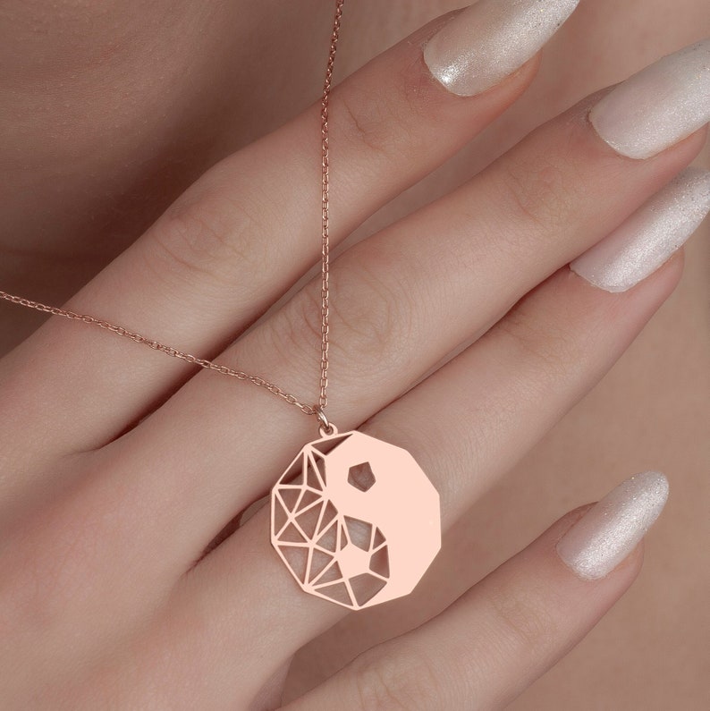 May include: A rose gold necklace with a geometric yin yang pendant. The pendant is a circle with a geometric design and a yin yang symbol in the center.