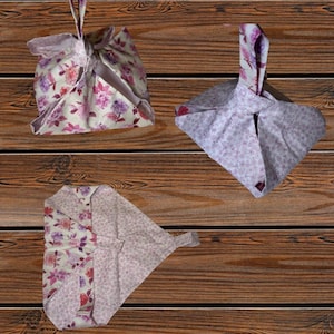 May include: Three fabric gift bags with floral patterns in shades of pink, purple, and white. The bags are folded and tied, with a handle at the top. The bags are displayed on a wooden surface.