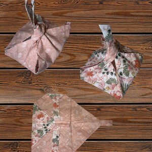 May include: Three fabric bags with floral patterns in pink, white, and green. The bags are folded and tied in different ways, showcasing their versatility. The background is a wooden surface.