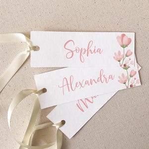 May include: White rectangular tags with the names "Sophia" and "Alexandra" in pink script. Each tag has a floral design in the upper right corner and a cream-colored ribbon. The tags are on a beige fabric surface.