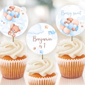 May include: Three cupcake toppers with a blue and brown balloon theme. One topper features a teddy bear holding a balloon, another says "Beary Sweet" and the last one says "Benjamin is 1".