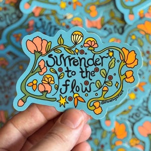 May include: A light blue sticker with the words "Surrender to the flow" in black script, surrounded by orange and yellow flowers and green leaves. The sticker has a floral design and the text "@floatinginplace".