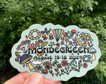 May include: A light blue sticker with a whimsical design featuring a variety of colorful flowers, mushrooms, and other nature-inspired elements. The text "Mondegreen" is written in a bold, stylized font, and the date "August 15-18 2024" is written below. The sticker has a cloud-like shape.