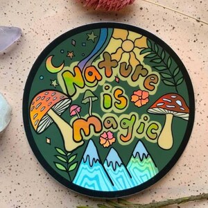 K&ouml;nnte beinhalten: Ein runder Aufkleber mit gr&uuml;nem Hintergrund und dem Text "Nature is magic" in Regenbogenfarben. Der Aufkleber zeigt Pilze, Berge, Sterne und eine Sonne.