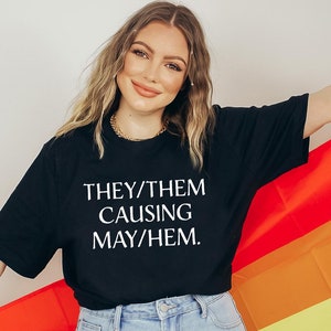 May include: A person wearing a black t-shirt with the text "THEY/THEM CAUSING MAY/HEM." The person is holding a rainbow pride flag.