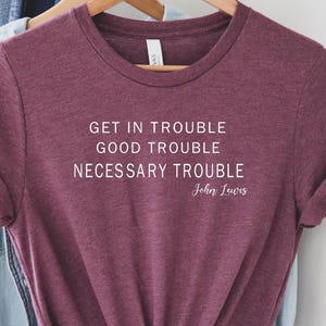 May include: A maroon short-sleeved t-shirt with white text that reads: GET IN TROUBLE GOOD TROUBLE NECESSARY TROUBLE John Lewis. The shirt features a crew neckline and is displayed on a wooden hanger, layered over a light blue denim shirt.