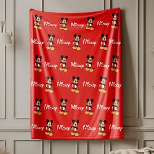 May include: A bright red blanket featuring a repeating pattern of Mickey Mouse illustrations and the white cursive text Mickey. The fabric displays the character in a classic pose with yellow shoes, white gloves, and red shorts against the solid red background.