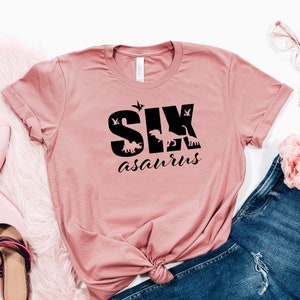 May include: A pink t-shirt with the text "SIX asaurus" in black. The text is surrounded by dinosaur silhouettes.
