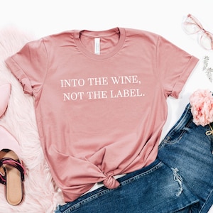 May include: A pink t-shirt with the text "INTO THE WINE, NOT THE LABEL." on it. The shirt is styled with blue jeans, pink shoes, and pink flowers.