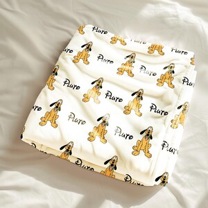 May include: A folded cream-colored blanket featuring a repeating pattern of the cartoon dog Pluto in a sitting position with his tongue out, interspersed with the word Pluto written in a black cursive script across the fabric surface.