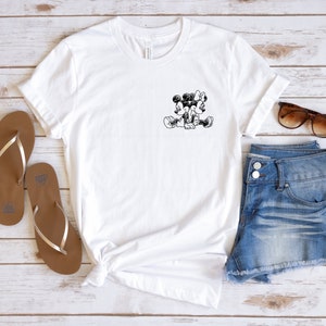 May include: White t-shirt with a black and white image of Mickey Mouse and Minnie Mouse holding hands. The t-shirt is laid out on a white wooden surface with a pair of brown flip-flops, denim shorts, and sunglasses.