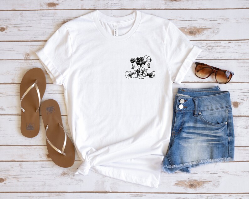 May include: White t-shirt with a black and white image of Mickey Mouse and Minnie Mouse holding hands. The t-shirt is laid out on a white wooden surface with a pair of brown flip-flops, denim shorts, and sunglasses.