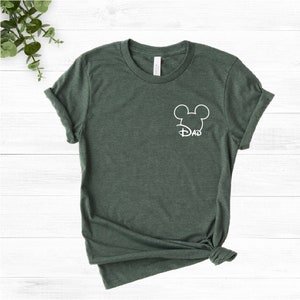 May include: Green t-shirt featuring a white Mickey Mouse outline and the word "DAD" underneath. The sleeves are rolled up, and the bottom is knotted. The shirt is displayed on a white wooden surface.