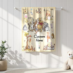 May include: A light yellow baby blanket featuring illustrations of Winnie the Pooh and friends. The blanket has a decorative border and includes the text "Custom Name". The blanket is hanging on a white rod against a white wall.