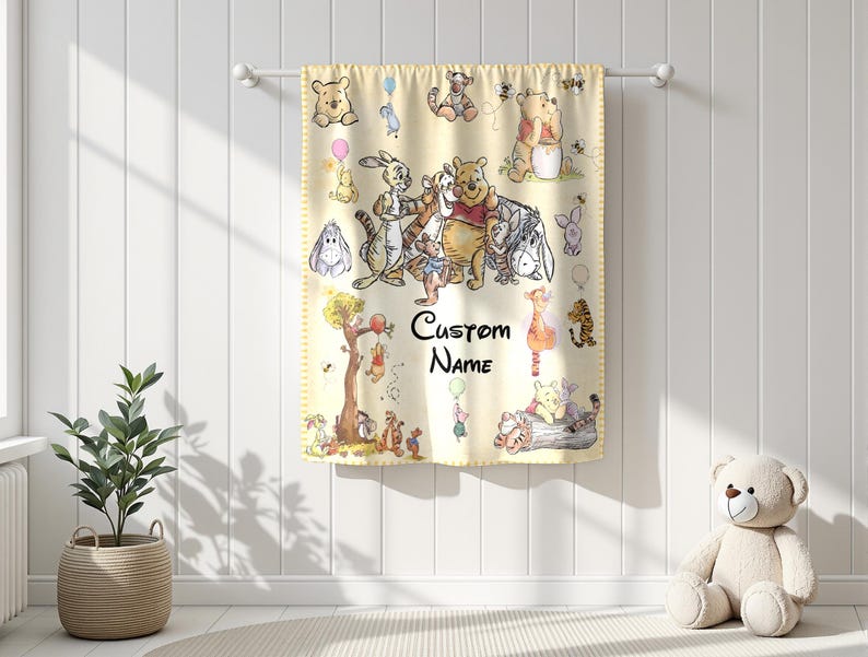 May include: A light yellow baby blanket featuring illustrations of Winnie the Pooh and friends. The blanket has a decorative border and includes the text "Custom Name". The blanket is hanging on a white rod against a white wall.