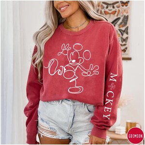 Puede incluir: Una sudadera roja de manga larga con un contorno blanco de Mickey Mouse y el texto "Mickey" en la manga.