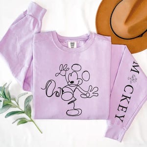 Puede incluir: Una sudadera de manga larga de color lavanda con un dibujo de Mickey Mouse en negro en la parte delantera y la palabra "Mickey" en una manga.