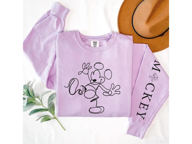 Puede incluir: Una sudadera de manga larga de color lavanda con un dibujo de Mickey Mouse en negro en la parte delantera y la palabra "Mickey" en una manga.