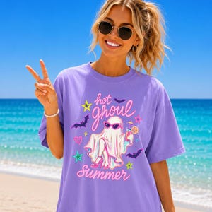 May include: A light purple short-sleeved t-shirt featuring a graphic print of a ghost wearing pink sunglasses and high heels, holding a cocktail. The text reads hot ghoul Summer, surrounded by colorful stars, bats, and a heart icon.