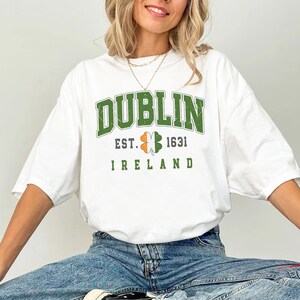 Peut inclure: T-shirt blanc avec un tr&egrave;fle graphique vert, blanc et orange et le texte "DUBLIN EST. 1631 IRLANDE".